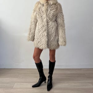 Pu&ograve; includere: Un cappotto di pelliccia colorata, lungo fino al ginocchio, con un motivo strutturato a forma di chevron. Il cappotto ha un colletto e maniche lunghe. La persona indossa stivali neri al ginocchio con punta e tacco piccolo.