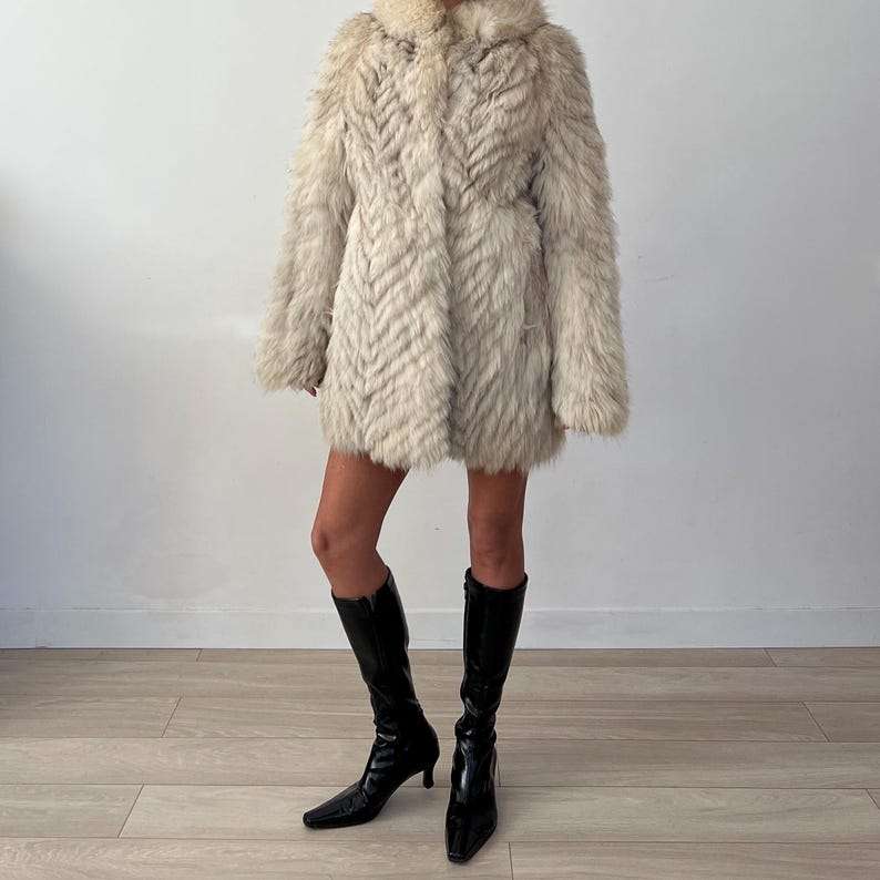 Pu&ograve; includere: Un cappotto di pelliccia colorata, lungo fino al ginocchio, con un motivo strutturato a forma di chevron. Il cappotto ha un colletto e maniche lunghe. La persona indossa stivali neri al ginocchio con punta e tacco piccolo.