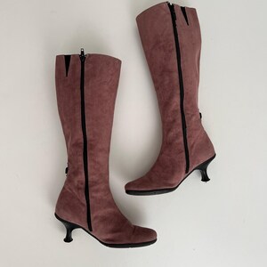 May include: A pair of mauve knee-high boots. The boots have a suede-like texture, black zippers, and small, curved heels. The boots are set against a white background.
