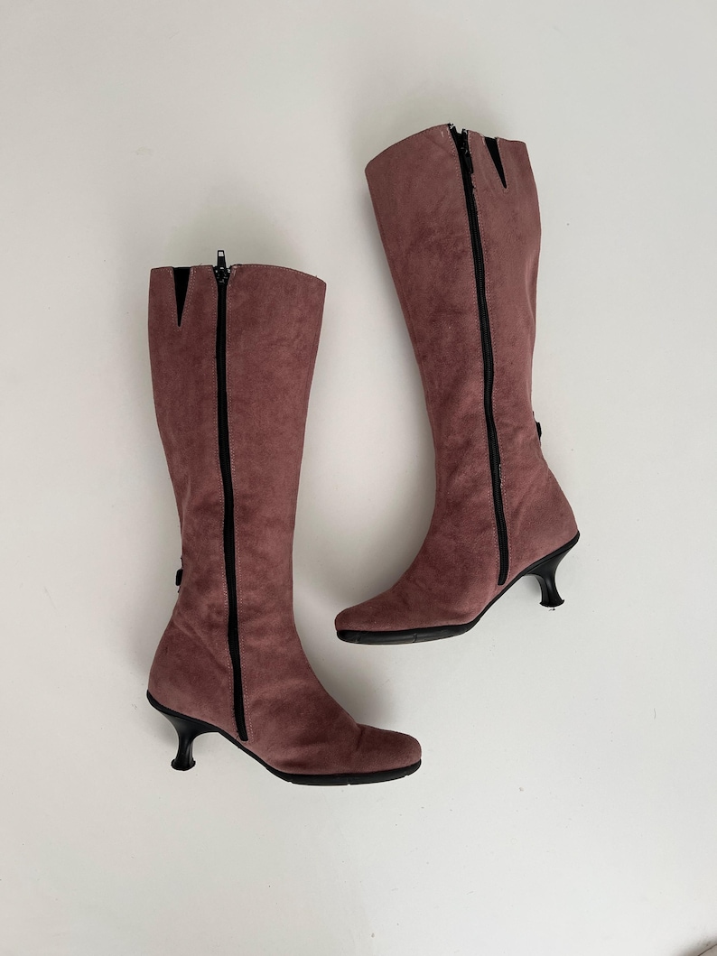 May include: A pair of mauve knee-high boots. The boots have a suede-like texture, black zippers, and small, curved heels. The boots are set against a white background.