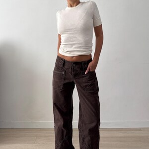 May include: A short-sleeved, cream-colored ribbed crewneck t-shirt paired with dark brown, low-rise cargo pants. The pants feature multiple pockets, including a zippered pocket on the right side, a button closure, and visible seam detailing.