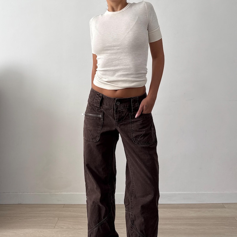 May include: A short-sleeved, cream-colored ribbed crewneck t-shirt paired with dark brown, low-rise cargo pants. The pants feature multiple pockets, including a zippered pocket on the right side, a button closure, and visible seam detailing.