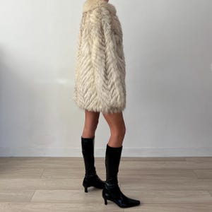 Pu&ograve; includere: Un cappotto di pelliccia color crema vintage con un motivo a spina di pesce. Il cappotto ha un colletto arrotondato e arriva a met&agrave; coscia. La persona indossa stivali neri al ginocchio con un piccolo tacco. Lo sfondo &egrave; una parete bianca e un pavimento in legno chiaro.