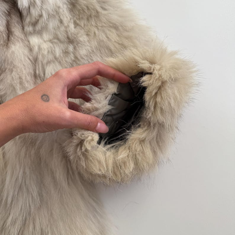 Pu&ograve; includere: Primo piano di una manica di cappotto di pelliccia beige chiaro. L'interno della manica rivela una fodera scura. La pelliccia &egrave; lunga e soffice, con un aspetto naturale e strutturato. Il cappotto sembra ben fatto e lussuoso.