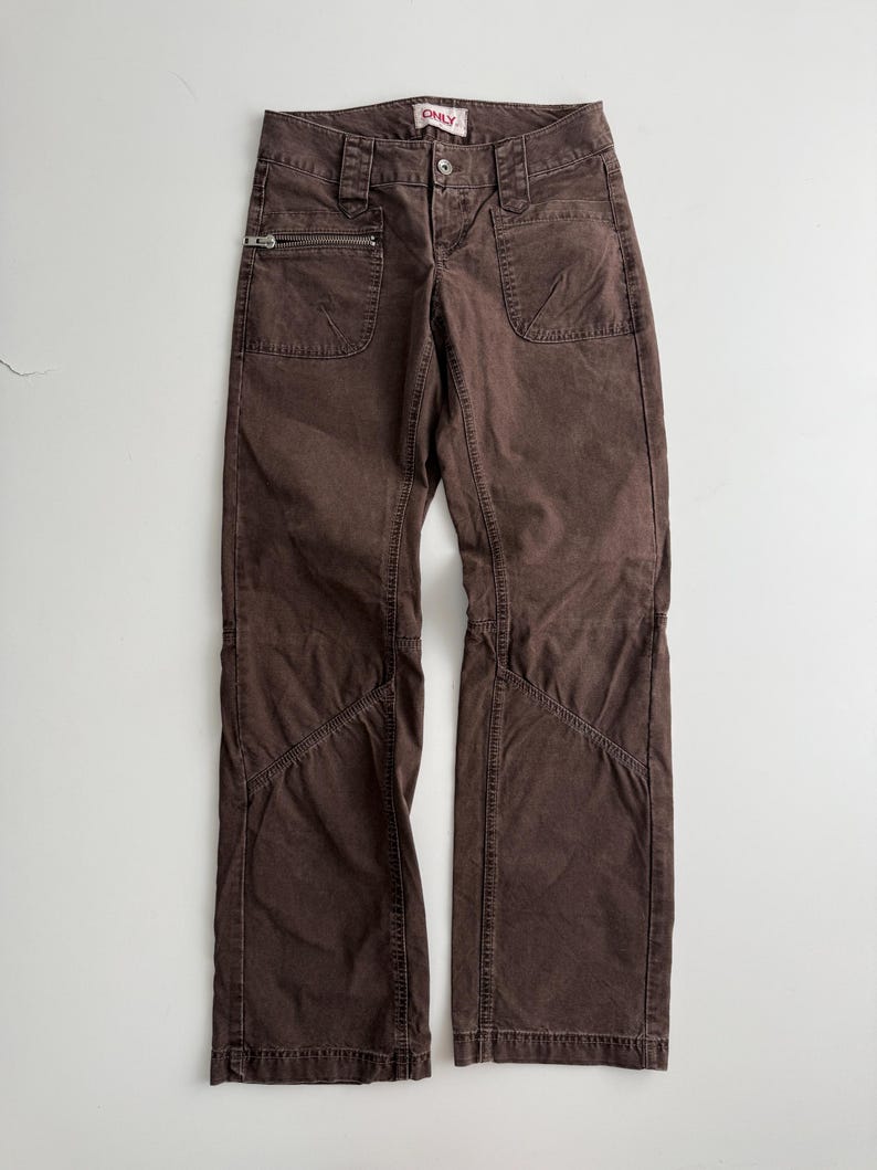 May include: Brown pants featuring a front zipper closure, a button at the waist, and two front pockets. One pocket includes a horizontal zipper detail. The pants have visible stitching along the legs and a label at the waistband that reads ONLY.