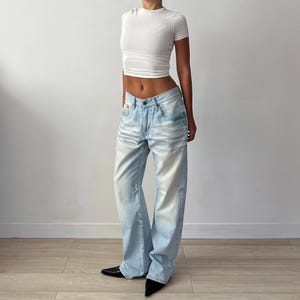 May include: Pale blue denim jeans with a faded wash and a wide leg. The jeans are worn with a white, short-sleeved, fitted top. The model is wearing black pointed-toe shoes.