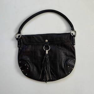 Könnte beinhalten: Eine schwarze Ledertasche mit Schlangenhautmuster und einem silbernen Quastenanhänger. Die Tasche hat einen Klappenverschluss und einen Henkel.