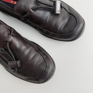 Puede incluir: Par de mocasines de cuero marr&oacute;n oscuro con punta redondeada. Cada zapato presenta un detalle met&aacute;lico decorativo y un detalle de cord&oacute;n negro. Los zapatos se muestran sobre una superficie blanca.