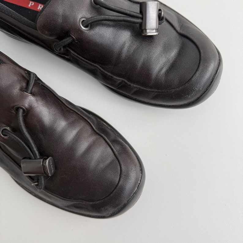 Puede incluir: Par de mocasines de cuero marr&oacute;n oscuro con punta redondeada. Cada zapato presenta un detalle met&aacute;lico decorativo y un detalle de cord&oacute;n negro. Los zapatos se muestran sobre una superficie blanca.