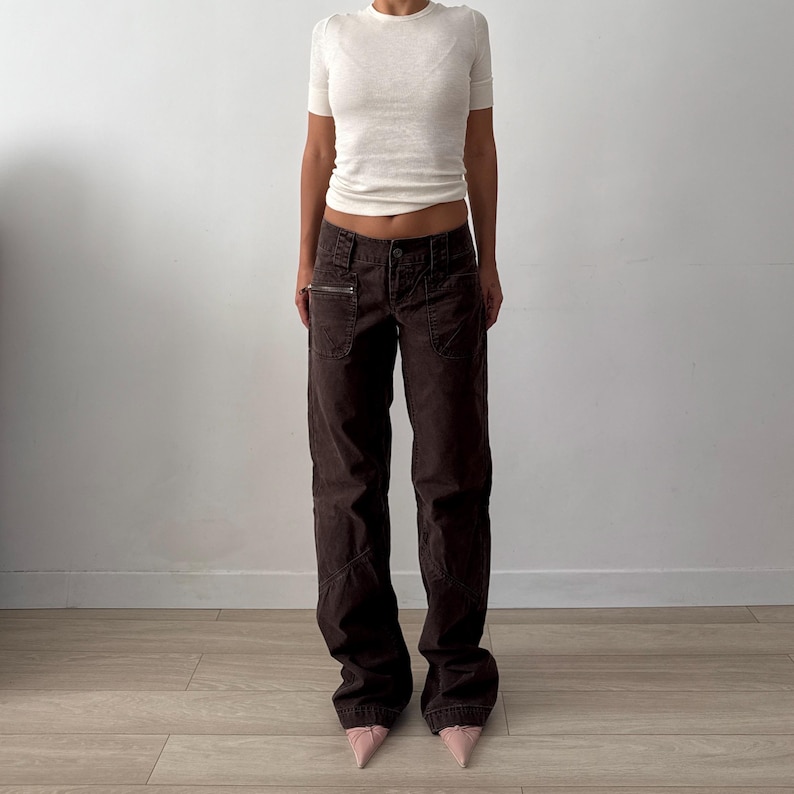 May include: A short-sleeved white crew neck t-shirt paired with dark brown low-rise cargo pants. The pants feature a button closure, belt loops, and a zippered pocket on the right side. The outfit is completed with light pink pointed-toe shoes.