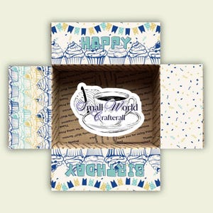 Può includere: Una scatola regalo blu e bianca con un design a cupcake e le parole "Happy Birthday". La scatola è aperta e all'interno c'è un adesivo che dice "Small World Crafterall" con una tazza di caffè su un piattino.