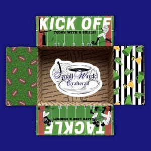 May include: A brown cardboard box with a green and white football field design. The box is open and the inside is lined with a green and white pattern of footballs. The top of the box has the words "Kick Off Today With A Smile!" in black text. The box also has a sticker with a cup of coffee and the words "Small World Crafterall".