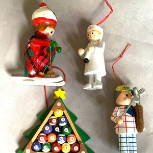 May include: A collection of Christmas ornaments featuring a skier, a doctor, a golfer, and a Christmas tree with billiard balls. The ornaments are made of wood and painted in various colors, with red string hangers.