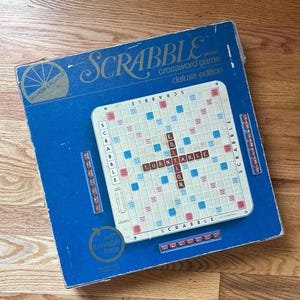 Pode incluir: Uma caixa azul do jogo de palavras cruzadas Scrabble edição deluxe com um tabuleiro branco com casas vermelhas e azuis. O tabuleiro exibe a palavra TURNTABLE no centro. A caixa inclui um logótipo circular e uma base giratória.