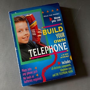 May include: A rectangular product box titled BUILD YOUR OWN TELEPHONE. The blue box features a graphic of a person holding a red telephone handset. Text includes HOLD THE PHONE! Now you can, Includes all electronic components, telephone case, and THE TELEPHONE BOOK, Plugs into any phone jack, No tools or batteries required.