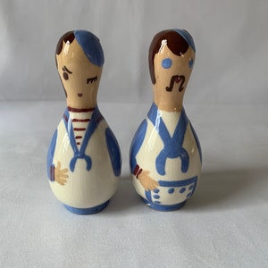 Op de afbeelding: Een paar vintage keramische zout- en peperstrooiers. Elke strooier heeft de vorm van een persoon met een blauwe matrozenhoed, blauw schort en wit overhemd. Eén heeft een rood-wit gestreept shirt en de ander een snor.