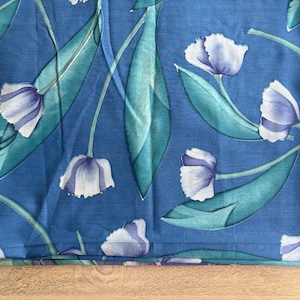 May include: A blue fabric featuring a repeating pattern of light purple and white tulip-like flowers with long, pointed teal green leaves. The floral design is spread across the surface, showcasing soft color gradients on the petals and foliage.