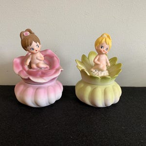 May include: Two ceramic figurines of nude babies, one in a pink flower and the other in a yellow flower. The figurines have painted facial features and are sitting atop decorative containers. The background is a neutral color.