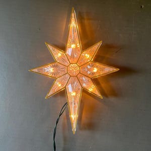 May include: A multi-pointed star-shaped light fitting featuring clear, faceted glass panels framed in gold-toned metal. The interior emits a warm, glowing light, and a thin black electrical cord extends from the bottom point of the star structure.