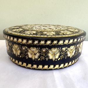 May include: A round black metal tin featuring a decorative floral pattern in cream, gold, and white. The lid and sides are adorned with intricate botanical illustrations and a repeating leaf border, creating a detailed and ornate appearance.