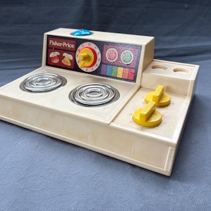 以下が含まれることがあります： シルバーのバーナーが付いた、オフホワイトのヴィンテージFisher-Priceおもちゃのストーブ。黄色のノブ、青いボタン、カラフルなディテールが施されたコントロールパネルが特徴です。Fisher-Priceのロゴが見えます。