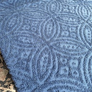 Könnte beinhalten: Eine blaue Textildecke mit einem erhabenen, getufteten und strukturierten geometrischen Muster aus sich wiederholenden Ovalen und Rautenformen. Der Stoff hat eine weiche, geschlungene Florstruktur, die ein dreidimensionales Design erzeugt.