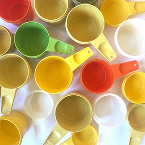 May include: A collection of plastic measuring cups in various colours including red, green, yellow, orange, and white. Each cup features a round bowl and a flat handle with a hole at the end. The cups are arranged in a scattered, overlapping pattern.
