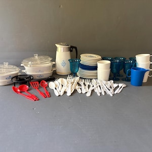May include: A collection of toy kitchenware including white lidded casserole dishes with blue floral patterns, a white pitcher, stacks of white and blue plates, blue goblets, white mugs, a blue pitcher, and various red and white plastic utensils.