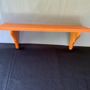 Könnte beinhalten: Ein Holzregal mit leuchtend orangefarbenem Finish. Das Regal hat eine rechteckige Oberseite und wird von zwei dekorativen Halterungen getragen. Die Halterungen sind aus hellerem Holz, was einen Kontrast erzeugt. Zur Wandmontage.