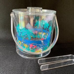 May include: A clear plastic ice bucket with a tropical print featuring palm trees, flowers, and a blue ocean. The bucket has two handles and comes with a pair of clear plastic tongs.