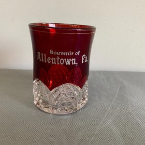 Könnte beinhalten: Ein rubinrotes Souvenirglas mit einem transparenten, geschliffenen Glasfuß. Auf dem Glas steht in weißer Schrift "Souvenir of Allentown, Pa.". Das Glas hat eine ausgestellte Form.