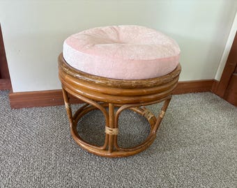May include: A round rattan stool with a light pink cushion. The stool has a woven rattan frame with curved legs and a circular seat. The cushion is a soft, plush material.