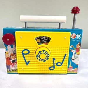 May include: A Fisher-Price toy radio with a blue body, a yellow speaker grille, and a white handle. It features a red side knob, a red antenna, and a central yellow dial labelled "TURN" with musical notes. Illustrations include "The Farmer in the Dell" text.