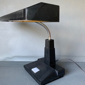 Op de afbeelding: Zwarte bureaulamp met een rechthoekige kap en een flexibele, zilverkleurige zwanenhalsarm. De lamp heeft een zwarte voet en een netsnoer. De lamp is ontworpen voor gebruik op een bureau en biedt gerichte verlichting.