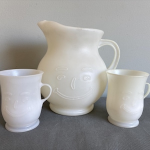 May include: A vintage white jug and two matching mugs. The jug and mugs feature a smiling face design. The jug has a handle and a spout. The mugs have handles and a rounded base. The set is made of a light-coloured plastic.