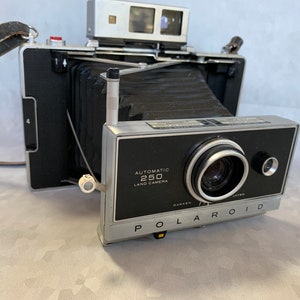 Op de afbeelding: Vintage Polaroid 250 Land Camera met een zwart en zilverkleurig lichaam, een leren riem en een zilveren flitser. De camera heeft een opvouwbare balg en de tekst "Automatic 250 Land Camera" staat op de voorkant.