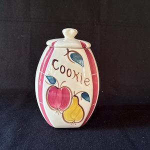 May include: A ceramic cookie jar with a lid. The jar is white with vertical pink stripes and features a hand-painted design of an apple and a pear. The word "Cookie" is written in brown script.