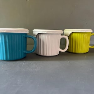 May include: Three ceramic mugs with vertical ribbed textures and matching white plastic lids. The mugs are colored in bright turquoise, solid white, and lime green. Each mug features a side handle and a cylindrical shape with a flat base.