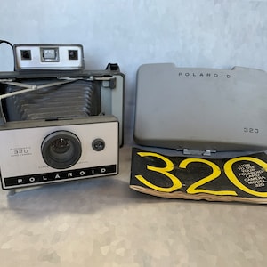 Op de afbeelding: Vintage Polaroid 320 Land Camera met een grijze behuizing en een zwarte riem. De camera heeft een wit en zwart voorpaneel met de tekst "Polaroid" en "Automatic 320 Land Camera". Een grijze hoes en een handleiding zijn ook inbegrepen.