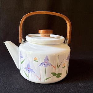 May include: White enamel teapot with a wooden handle and lid. The teapot is decorated with purple iris flowers, green leaves, and yellow accents. The handle is arched and made of light brown wood.