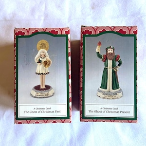 May include: Two decorative figurines from "A Christmas Carol" in their original boxes. One figure is a woman in a white dress, the other is a man in a green robe. The boxes have a red and green design.