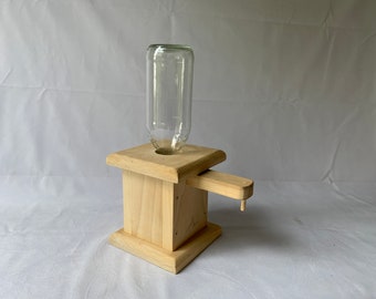 May include: A wooden soap dispenser with a clear glass bottle inserted at the top. The dispenser has a square base and a spout. The wood is light-colored and appears unfinished.