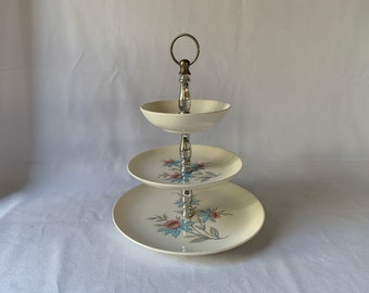 May include: A three-tiered serving stand with a cream-colored finish. Each tier is a different size, with a floral design in blue and pink. The stand has a silver-colored metal handle and support.