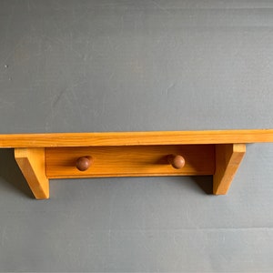 May include: A light-coloured wooden shelf with two wooden knobs below the shelf. The shelf is mounted on a grey wall. The shelf is made of light-coloured wood.