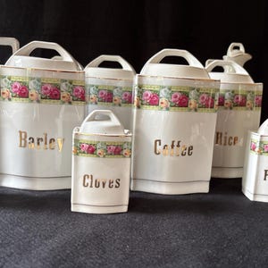 Op de afbeelding: Een verzameling witte keramische keukencanners met bloemenrand en gouden letters. De set bevat potten met de labels "Gerst", "Koffie", "Rijst", "Peper", "Nootmuskaat" en "Kruidnagel", plus een melkkan.