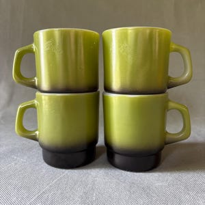 May include: Four green and black mugs stacked in a square formation. The mugs have a rounded handle and a two-tone color scheme, with a lighter green top and a dark black base. The mugs are made of ceramic.