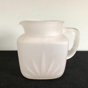 Könnte beinhalten: Eine Milchkanne aus mattiertem Glas mit Griff und Ausguss. Die Kanne hat einen quadratischen Boden mit einem Sonnenstrahl-Design und eine abgerundete Oberseite. Die Kanne steht auf einer schwarzen Oberfläche.