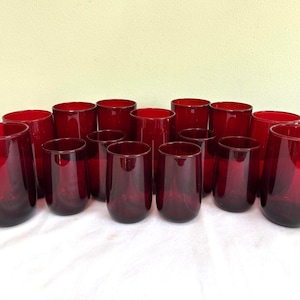 May include: A collection of fourteen cylindrical drinking glasses made of translucent red glass. The glasses are arranged in a group, featuring smooth, rounded bases and straight sides, creating a uniform set of glassware for serving beverages.