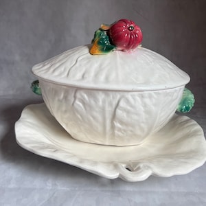 May include: Cream-colored ceramic soup tureen with a matching leaf-shaped platter. The tureen lid is topped with a red tomato and green leaves. The tureen and platter have a textured, cabbage-like design. Handles on the tureen are green.