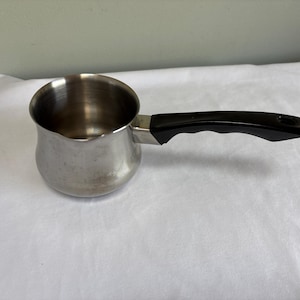 May include: A stainless steel butter warmer or small melting pot with a rounded base and a single black plastic handle. The pot features a polished metallic finish, a wide circular opening, and a contoured handle with a hole at the end.